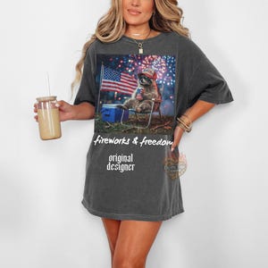 May include: Dark gray t-shirt featuring a graphic of a raccoon wearing a patriotic hat, sitting in a chair, with an American flag and fireworks in the background. The text "fireworks & freedom" and "original designer" are printed on the shirt.