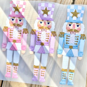 May include: Three flat nutcracker figures in pink, lavender, and light blue. Each figure wears a checkered hat with a gold glitter star, a matching uniform with gold glitter accents on the shoulders, belt, and boots, and has a painted face.