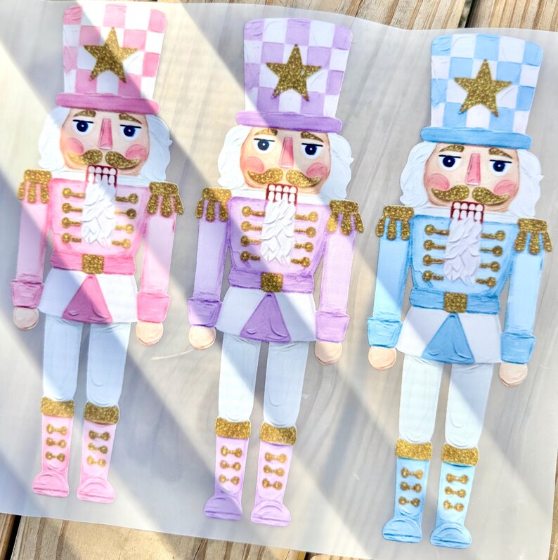 May include: Three flat nutcracker figures in pink, lavender, and light blue. Each figure wears a checkered hat with a gold glitter star, a matching uniform with gold glitter accents on the shoulders, belt, and boots, and has a painted face.