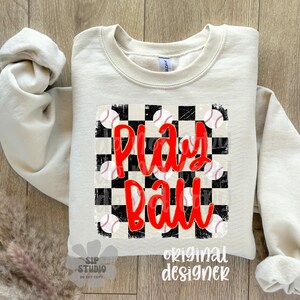 May include: A cream-colored sweatshirt with the words "Play Ball" in red script over a black and white checkered design. Baseball graphics are scattered around the text. The sleeves are rolled up.