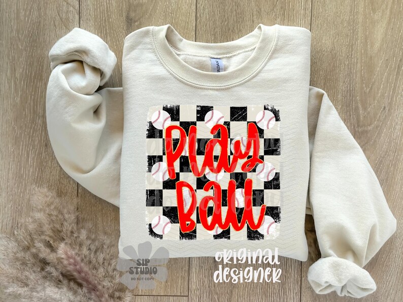 May include: A cream-colored sweatshirt with the words "Play Ball" in red script over a black and white checkered design. Baseball graphics are scattered around the text. The sleeves are rolled up.