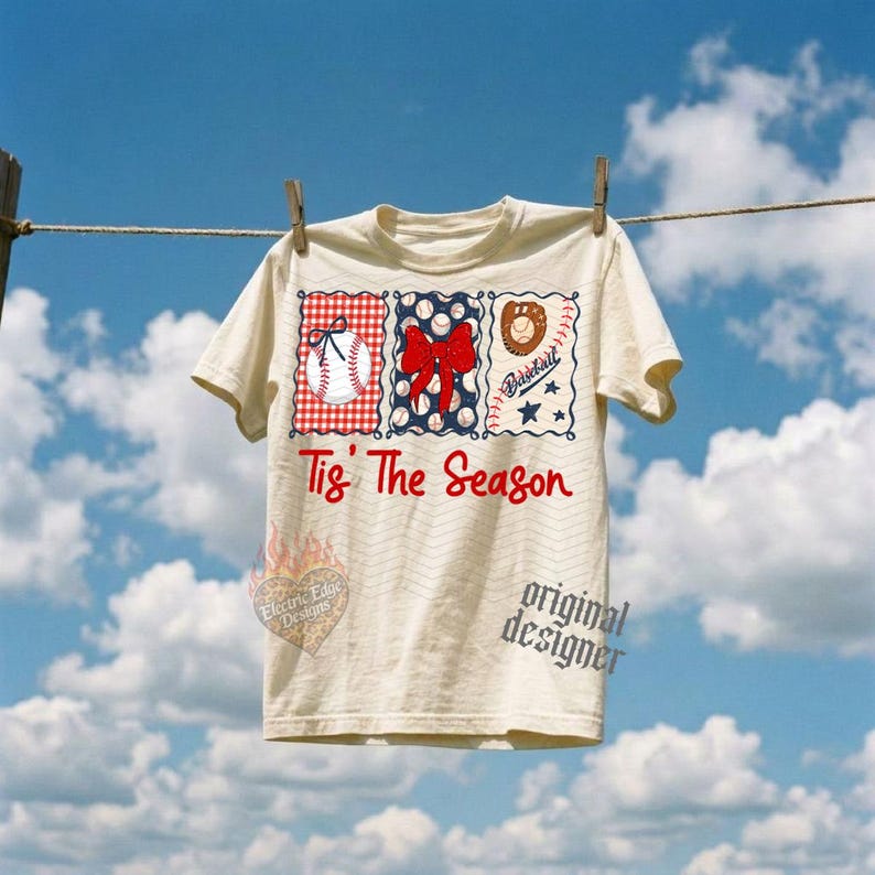 May include: A cream-colored t-shirt featuring three rectangular panels with baseball-themed graphics, including a baseball, a red bow, and a glove. The text 'Tis' The Season' is printed in red script below the panels. The shirt is hanging on a line.