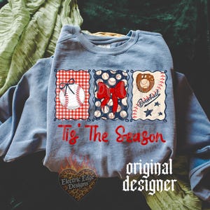 May include: A light blue crewneck sweatshirt featuring a graphic with three rectangular panels. The panels display a baseball with a bow, a red bow over baseballs, and a baseball glove with the text 'Tis The Season' in red script below.