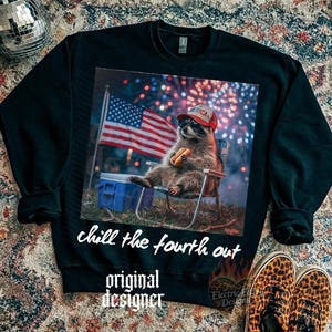 K&ouml;nnte beinhalten: Schwarzes Sweatshirt mit einem Aufdruck eines Waschb&auml;ren, der einen roten Hut mit dem Wort "America" tr&auml;gt und einen Hotdog h&auml;lt. Der Waschb&auml;r sitzt in einem Stuhl vor einer amerikanischen Flagge und Feuerwerk. Der Text "chill the fourth out" und "original designer" sind ebenfalls vorhanden.