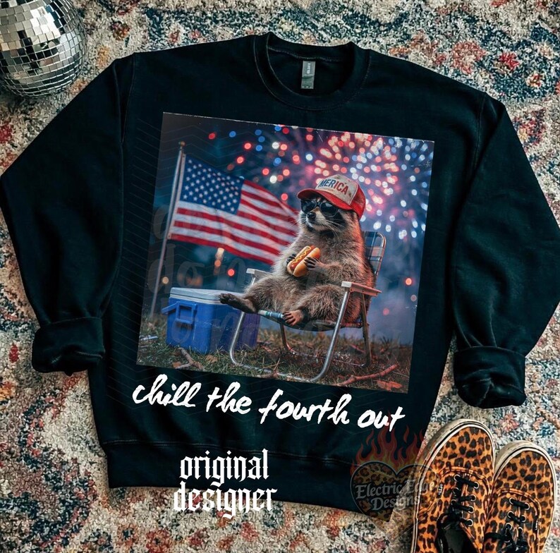 K&ouml;nnte beinhalten: Schwarzes Sweatshirt mit einem Aufdruck eines Waschb&auml;ren, der einen roten Hut mit dem Wort "America" tr&auml;gt und einen Hotdog h&auml;lt. Der Waschb&auml;r sitzt in einem Stuhl vor einer amerikanischen Flagge und Feuerwerk. Der Text "chill the fourth out" und "original designer" sind ebenfalls vorhanden.