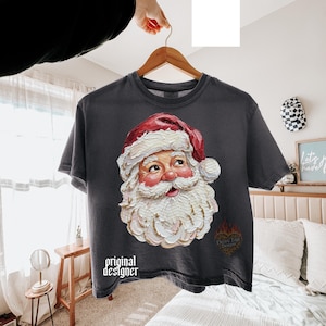 Puede incluir: Una camiseta de manga corta gris carb&oacute;n con un gr&aacute;fico de estilo pintura al &oacute;leo texturizada de Santa Claus con gorro rojo y barba blanca. El texto "original designer" est&aacute; impreso en blanco en la parte inferior izquierda, con un logo de coraz&oacute;n.