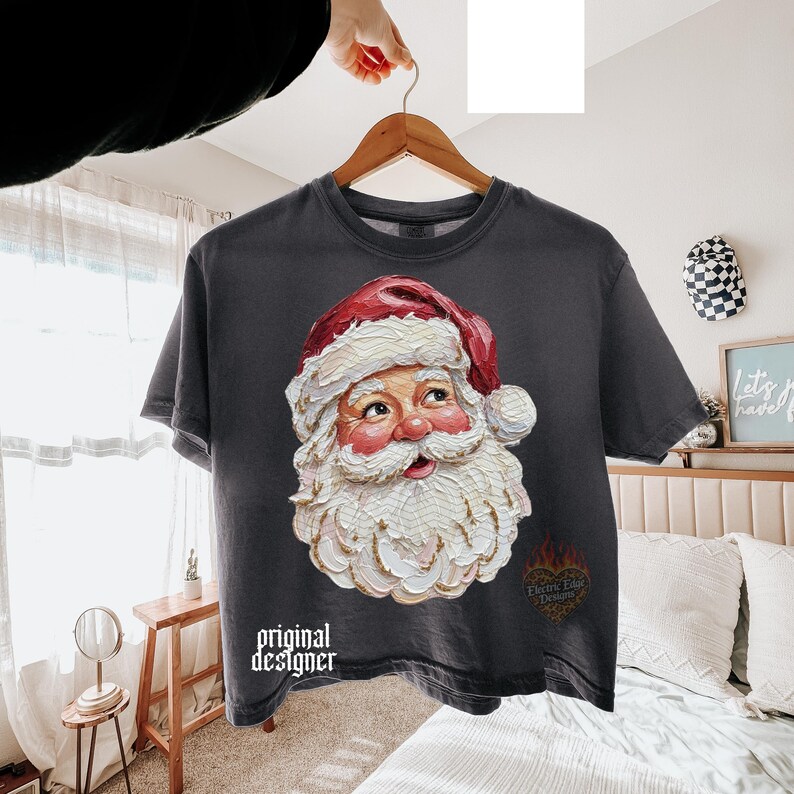 Puede incluir: Una camiseta de manga corta gris carb&oacute;n con un gr&aacute;fico de estilo pintura al &oacute;leo texturizada de Santa Claus con gorro rojo y barba blanca. El texto "original designer" est&aacute; impreso en blanco en la parte inferior izquierda, con un logo de coraz&oacute;n.