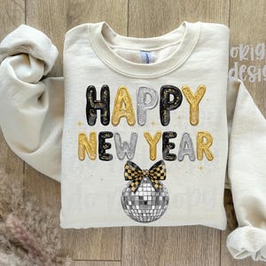 May include: A cream-coloured sweatshirt featuring the text "HAPPY NEW YEAR" in gold, silver, and black bubble letters. A disco ball and bow tie graphic are below the text. The sleeves are rolled up.