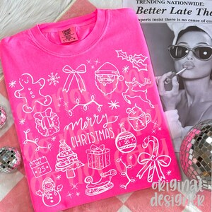 May include: A bright pink crew neck sweatshirt with a white Christmas-themed design. The design includes a gingerbread man, Santa, reindeer, Christmas tree, and the words "Merry Christmas".