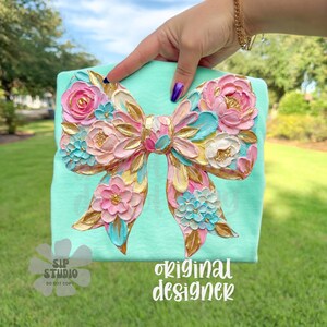 May include: A mint green t-shirt featuring a large, textured bow design. The bow is adorned with pink, blue, and white flowers, accented with gold details. The text "original designer" is visible on the shirt.
