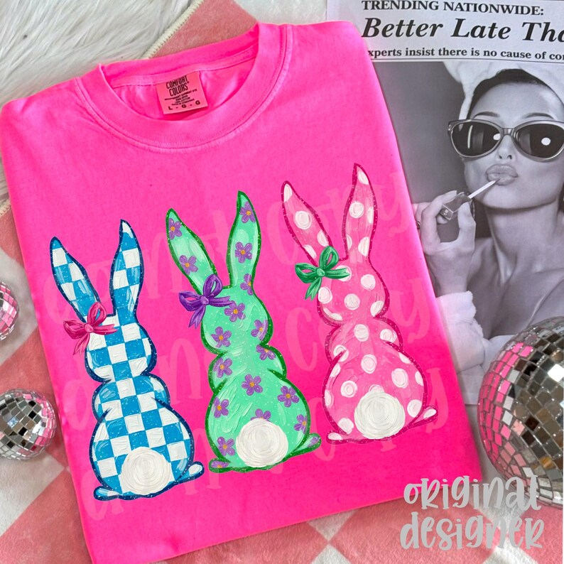 May include: Bright pink Comfort Colors sweatshirt featuring three Easter bunny designs. The bunnies are in blue and white checkered, green with purple flowers, and pink with white polka dots. Each bunny has a bow. The words "original designer" are at the bottom.