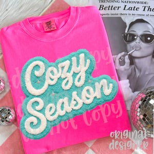 May include: A bright pink sweatshirt with the words "Cozy Season" in a white, textured font over a turquoise background. The sweatshirt has a crew neck and long sleeves. The text "Comfort Colors" is visible on the tag.