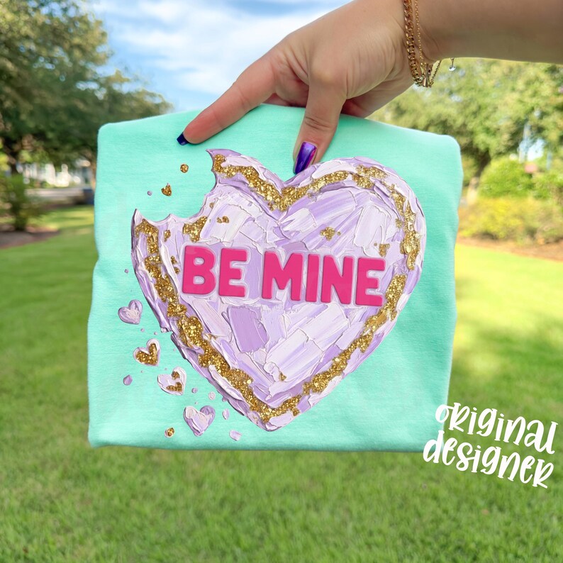 Op de afbeelding: Een mintgroen t-shirt met een gestructureerd hartontwerp. Het hart is een mix van paars en wit met gouden glitteraccenten en de woorden "BE MINE" in roze. Kleine hartdetails zijn verspreid rond het hoofdontwerp.