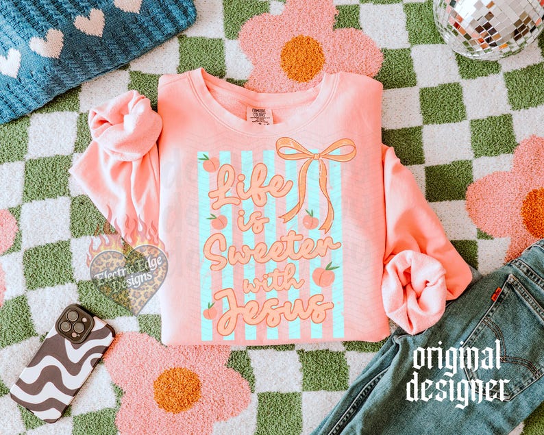 May include: A coral-colored sweatshirt with the text "Life is Sweeter with Jesus" in a playful font, accented with peaches and a bow. The design is set against light blue stripes. The sweatshirt is displayed on a checkered green and white surface.
