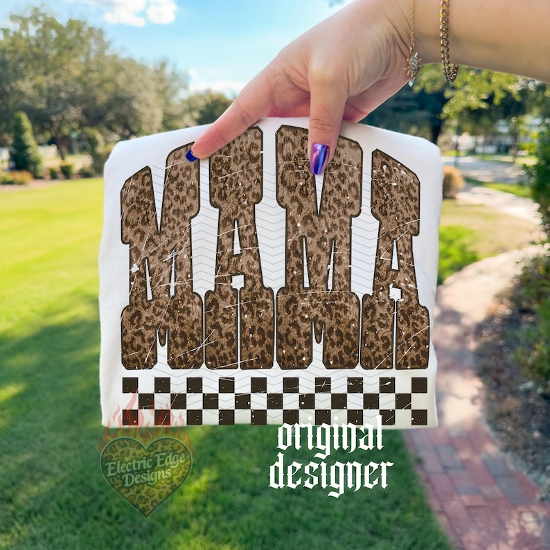 May include: White t-shirt with the word "MAMA" in brown leopard print lettering. The shirt has a black and white checkered pattern at the bottom and the words "original designer" in a gothic font.