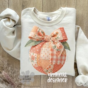 Op de afbeelding: Cr&egrave;mekleurige sweatshirt met een perzikontwerp, een strik en bladeren. De perzik is gemaakt van patchworkstof met bloemen- en polkadotpatronen. De woorden "original designer" staan rechtsonder gedrukt.