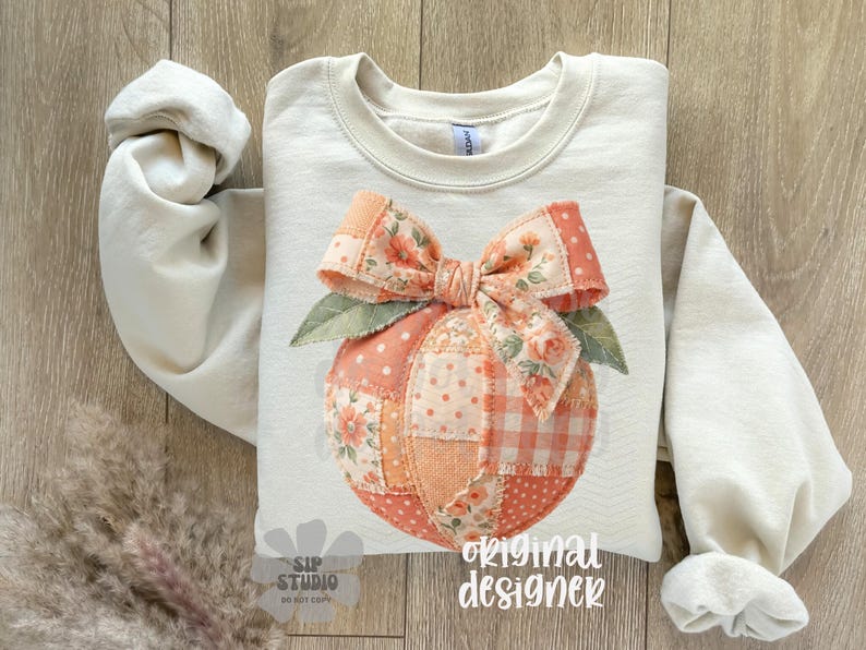 Op de afbeelding: Cr&egrave;mekleurige sweatshirt met een perzikontwerp, een strik en bladeren. De perzik is gemaakt van patchworkstof met bloemen- en polkadotpatronen. De woorden "original designer" staan rechtsonder gedrukt.