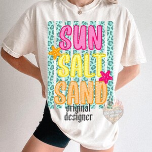 May include: Cream-colored t-shirt with the words "SUN SALT SAND" in pink, yellow, and orange, with starfish accents. The design is set against a teal leopard print background. The words "original designer" are below the main text.