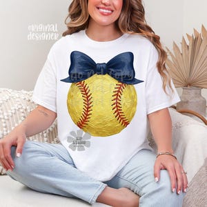 Pu&ograve; includere: T-shirt bianca con un disegno di softball giallo, cuciture rosse e un fiocco blu navy. La maglietta ha il testo "original designer" in alto a sinistra. L'immagine contiene anche il testo "do not copy" e "SIP studio".