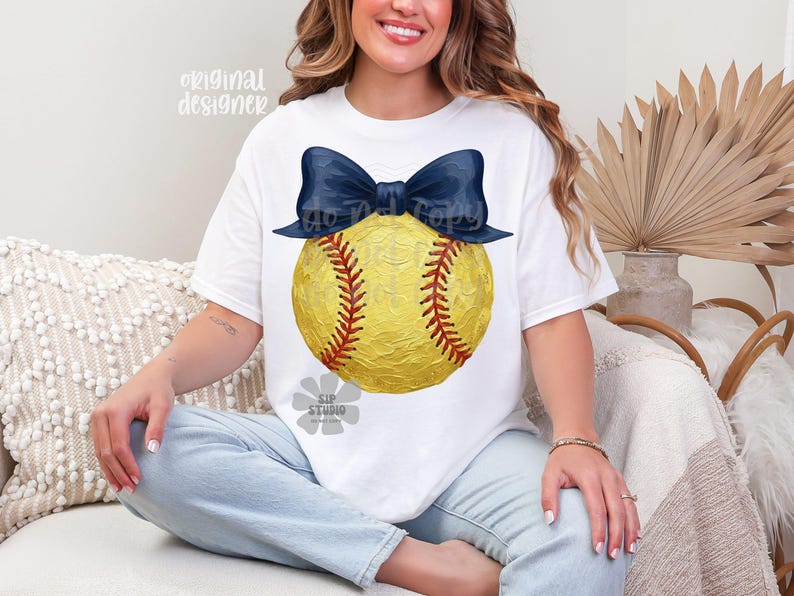 Pu&ograve; includere: T-shirt bianca con un disegno di softball giallo, cuciture rosse e un fiocco blu navy. La maglietta ha il testo "original designer" in alto a sinistra. L'immagine contiene anche il testo "do not copy" e "SIP studio".