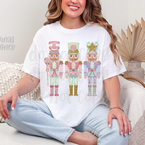 May include: White t-shirt featuring three pastel-colored nutcracker soldier illustrations. Each nutcracker has a unique design, with pink, green, and blue accents. The text "original designer" is visible on the shirt.