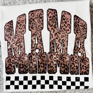 May include: Cream-colored t-shirt with the word "MAMA" in large, distressed, leopard print letters. A black and white checkered pattern borders the bottom of the shirt.