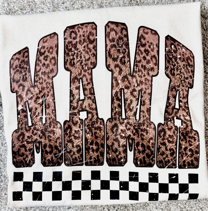 May include: Cream-colored t-shirt with the word "MAMA" in large, distressed, leopard print letters. A black and white checkered pattern borders the bottom of the shirt.