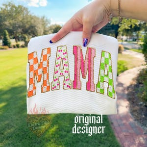 May include: White t-shirt with the word "MAMA" in colorful, textured letters. The "M" is orange and white checkered, the first "A" is cherry-patterned, the second "M" is pink, and the last "A" is green and white checkered. Gold glitter outlines the letters.