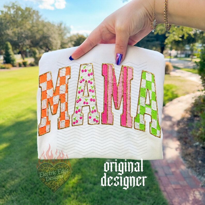 May include: White t-shirt with the word "MAMA" in colorful, textured letters. The "M" is orange and white checkered, the first "A" is cherry-patterned, the second "M" is pink, and the last "A" is green and white checkered. Gold glitter outlines the letters.
