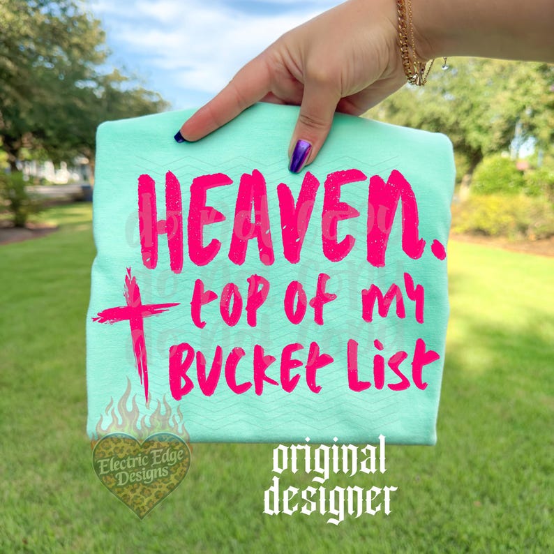May include: A light teal t-shirt with the words "HEAVEN. top of my BUCKET List" in bright pink. A pink cross is below "HEAVEN." The shirt also features a logo and the words "original designer" in white.