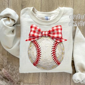 Peut inclure: Sweat-shirt cr&egrave;me avec un motif de baseball. La balle a des coutures rouges et est surmont&eacute;e d'un n&oelig;ud vichy rouge et blanc. Les mots "original designer" sont imprim&eacute;s en haut &agrave; droite.