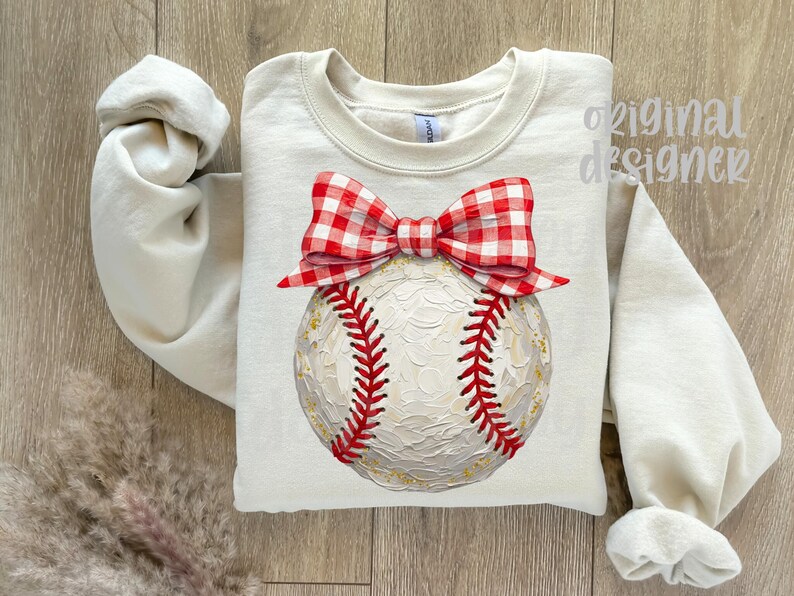 Peut inclure: Sweat-shirt cr&egrave;me avec un motif de baseball. La balle a des coutures rouges et est surmont&eacute;e d'un n&oelig;ud vichy rouge et blanc. Les mots "original designer" sont imprim&eacute;s en haut &agrave; droite.