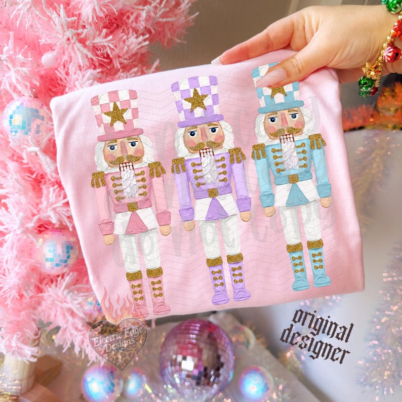 May include: A light pink t-shirt featuring a graphic print of three nutcracker figures in pastel pink, lavender, and light blue uniforms with gold glitter accents. Each nutcracker wears a tall hat with a gold star and stands in a rigid pose.