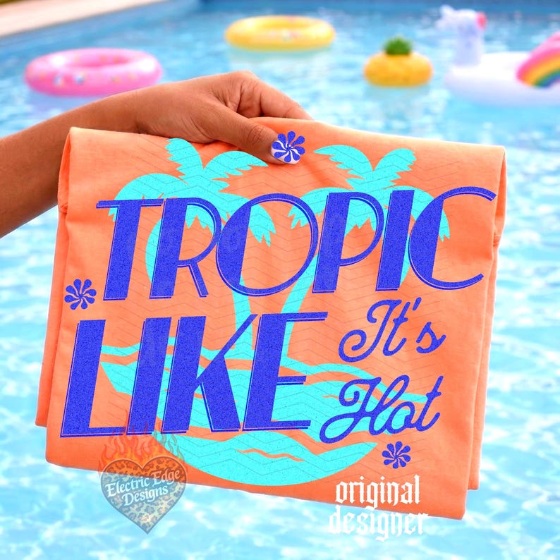 May include: An orange t-shirt featuring a large graphic with the text TROPIC LIKE It's Hot in bold blue letters. The design includes light blue palm tree silhouettes and small flower icons. A small heart-shaped logo with the text Electric Edge Designs is visible at the bottom left corner.