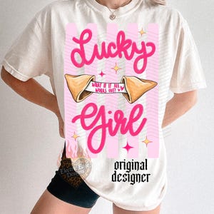 May include: Off-white t-shirt featuring a pink graphic with the words "Lucky Girl." The design includes a fortune cookie with the text "What if it all works out?" and star accents. The words "original designer" are also printed on the shirt.