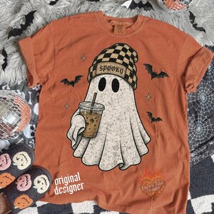 May include: A burnt orange t-shirt featuring a graphic of a ghost wearing a black and tan checkered beanie with the word SPOOKY printed on it. The ghost holds a coffee cup. Small black bats and stars surround the ghost. A heart logo is at the bottom.