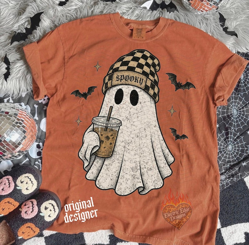 May include: A burnt orange t-shirt featuring a graphic of a ghost wearing a black and tan checkered beanie with the word SPOOKY printed on it. The ghost holds a coffee cup. Small black bats and stars surround the ghost. A heart logo is at the bottom.