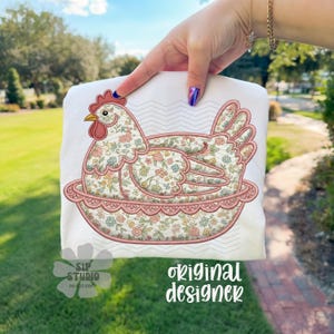 May include: White t-shirt featuring an embroidered hen design. The hen is in a nest, with floral patterns in pink, yellow, and blue. The design includes the text "original designer" and "SIP STUDIO DO NOT COPY".