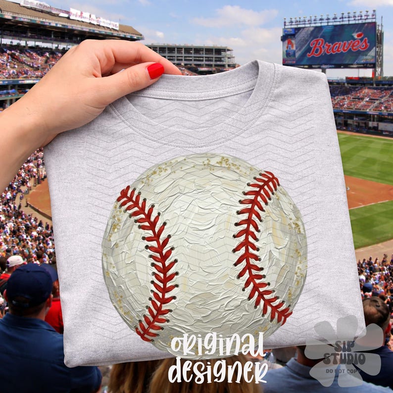 May include: A pale grey t-shirt with a large baseball design, showing red stitching. The words "original designer" are printed in a script font at the bottom. The shirt is held up in front of a baseball stadium with a large screen.