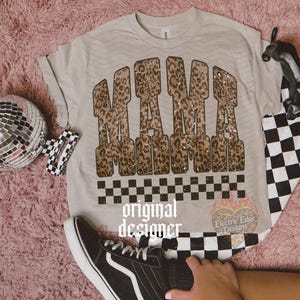 May include: Beige t-shirt with the word "MAMA" in a leopard print design. The shirt has a black and white checkered pattern at the bottom. A black and white checkered pattern is also visible on the right side of the shirt. A black sneaker is in the foreground.