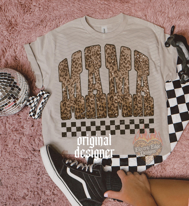 May include: Beige t-shirt with the word "MAMA" in a leopard print design. The shirt has a black and white checkered pattern at the bottom. A black and white checkered pattern is also visible on the right side of the shirt. A black sneaker is in the foreground.