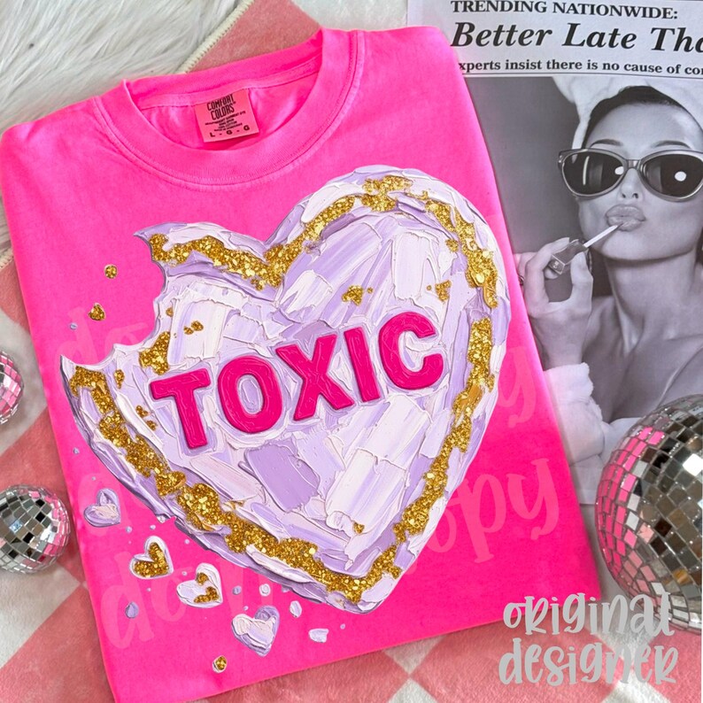 May include: A bright pink sweatshirt with a large, heart-shaped graphic that reads "TOXIC" in pink letters. The heart is a light purple color with gold glitter accents. The sweatshirt has a crew neck and long sleeves.