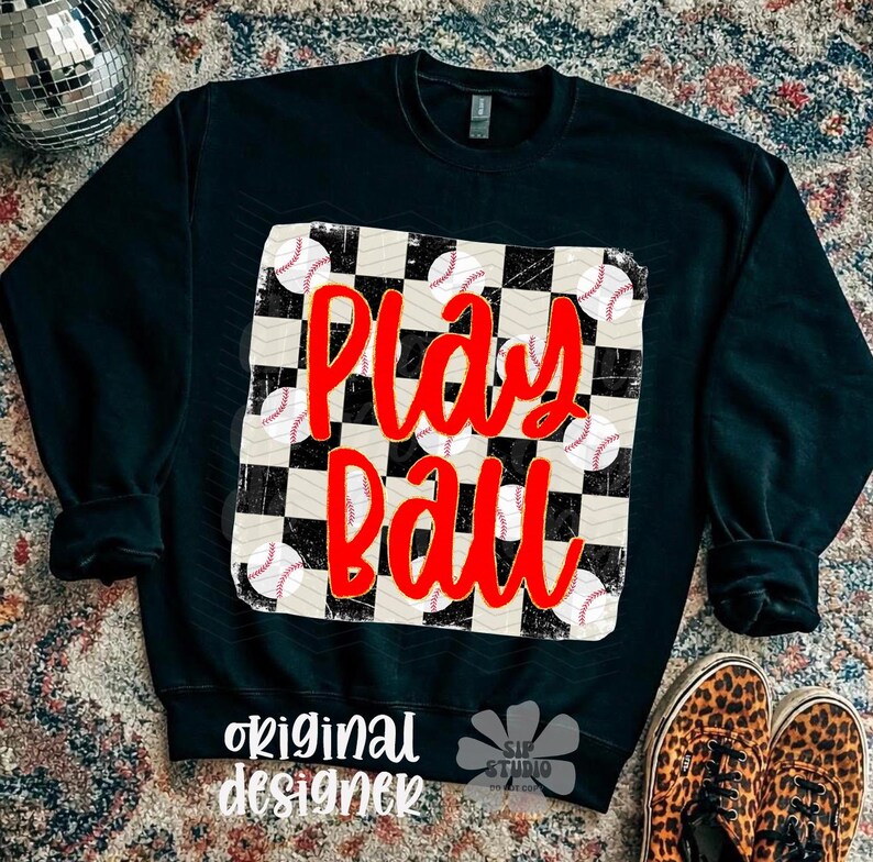 May include: Black sweatshirt with a baseball-themed design. The design features a checkered pattern with baseballs and the words "Play Ball" in red. The words "original designer" are printed at the bottom.