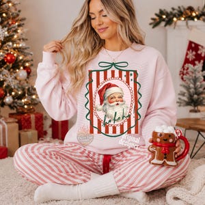 May include: A light pink crewneck sweatshirt featuring a central graphic of a vintage-style Santa Claus inside a red and white striped rectangular frame with a green bow at the top and the text ho ho ho written in cursive script below the face.