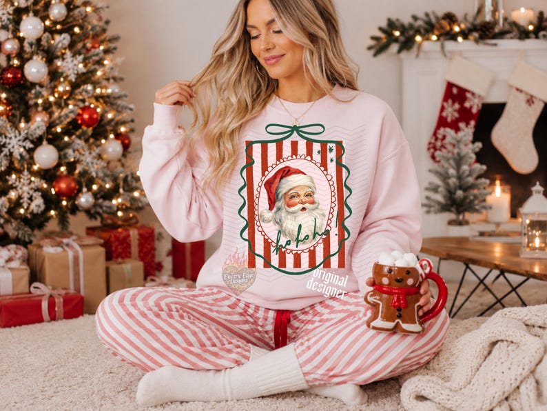 May include: A light pink crewneck sweatshirt featuring a central graphic of a vintage-style Santa Claus inside a red and white striped rectangular frame with a green bow at the top and the text ho ho ho written in cursive script below the face.