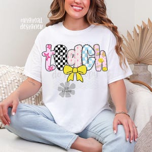 May include: White t-shirt with the word "TEACH" in a colorful, patterned design. The letters are decorated with stars, checkered patterns, and smiley faces. A yellow bow is centered below the word. The shirt is a casual style.