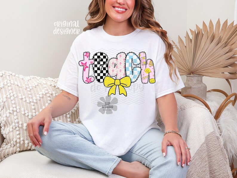 May include: White t-shirt with the word "TEACH" in a colorful, patterned design. The letters are decorated with stars, checkered patterns, and smiley faces. A yellow bow is centered below the word. The shirt is a casual style.