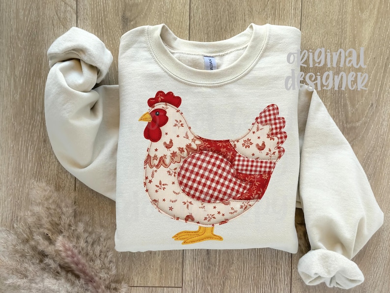 May include: A cream-coloured sweatshirt with a large, decorative chicken design. The chicken has a red comb and wattle, a yellow beak and feet, and a patchwork of red and white patterned fabrics. The words "original designer" are visible.