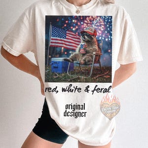 May include: Cream-colored t-shirt featuring a graphic of a raccoon wearing a red hat and sunglasses, seated in a chair with an American flag and fireworks in the background. Text reads "red, white & feral" and "original designer".