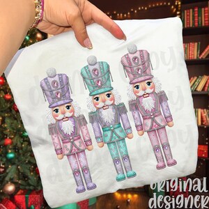 May include: A white cushion with three illustrated nutcracker soldiers in purple, teal, and pink. The soldiers have detailed uniforms and hats with decorative accents. The cushion is held by a hand. The design is festive and colourful.